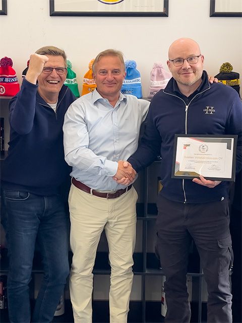 Kolme hymyilevää henkilöä poseeraa kameralle. Kaksi henkilöä kättelee, toisella on kädessään sertifikaatti.