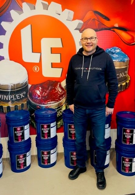 Tommi Uusi-Rauva poseeraa Lubrication Engineers -seinän edessä. Lattialla on Lubrication Engineersin rasvoja sinisissä muoviastioissa.